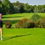 Woman playing golf on a vibrant green course with scenic tree backdrop.