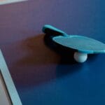 Close-up of a table tennis paddle and ball on a blue table, indoor setting.