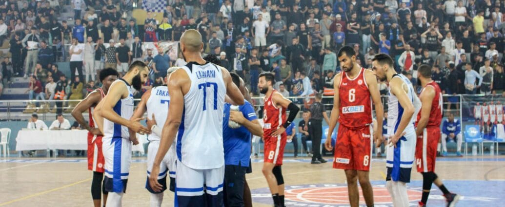 Basketball players in action during a competitive match with a full stadium crowd.