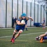 Dynamic rugby action shot featuring a player running with the ball at a match in Manchester, England.