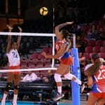 Dynamic action shot from a women's volleyball match with players in mid-air at the net.