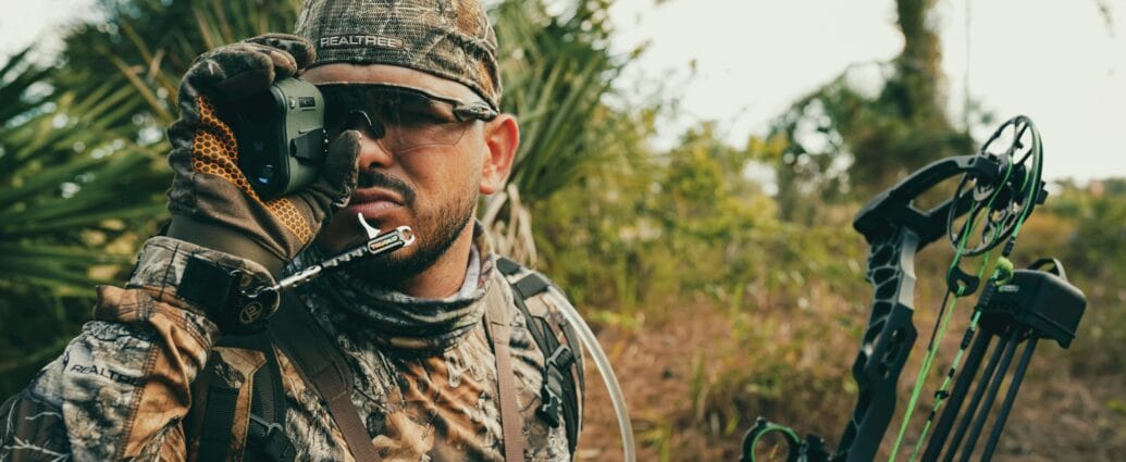 Hunter in camouflage gear using a rangefinder while outdoors, showcasing focus and precision.