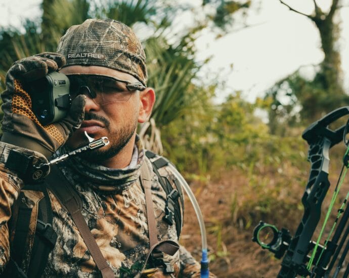 Hunter in camouflage gear using a rangefinder while outdoors, showcasing focus and precision.