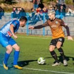 Two male soccer players in action during a competitive match on a grass field.