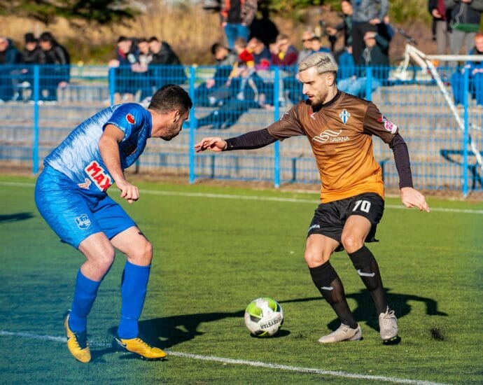 Two male soccer players in action during a competitive match on a grass field.