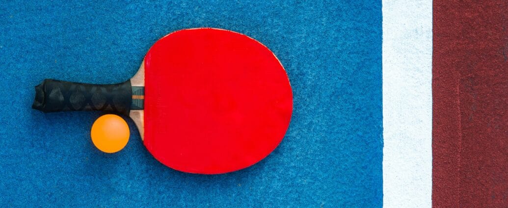 A red table tennis paddle and orange ball on a colorful court background.