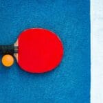 A red table tennis paddle and orange ball on a colorful court background.