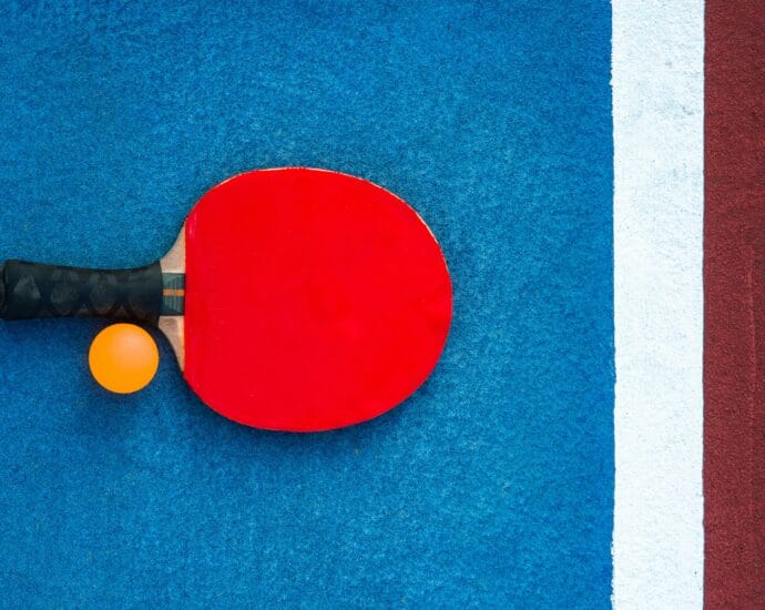 A red table tennis paddle and orange ball on a colorful court background.