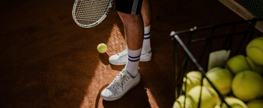 A tennis player stands on a clay court, showing sneakers, racket, and tennis balls in sunlight.