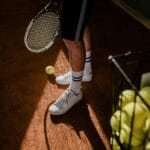 A tennis player stands on a clay court, showing sneakers, racket, and tennis balls in sunlight.