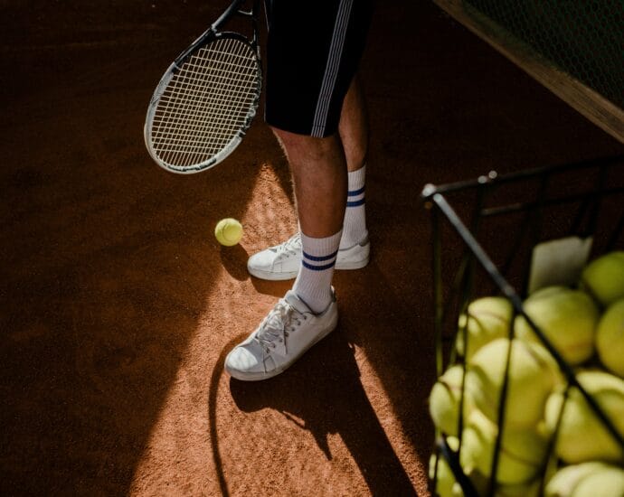 A tennis player stands on a clay court, showing sneakers, racket, and tennis balls in sunlight.