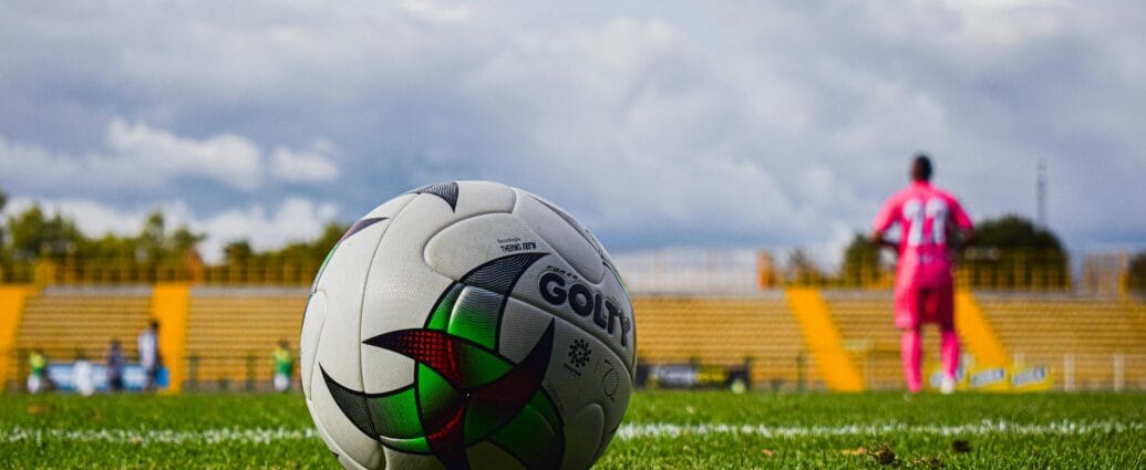 Close-up of a soccer ball on green grass field with a blurred player in pink jersey in the background during a match.