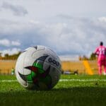 Close-up of a soccer ball on green grass field with a blurred player in pink jersey in the background during a match.