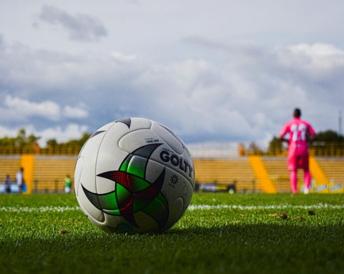 Close-up of a soccer ball on green grass field with a blurred player in pink jersey in the background during a match.