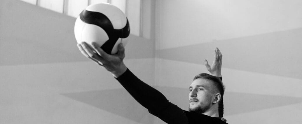 Caucasian man serving a volleyball in an indoor game setting, black and white photo.