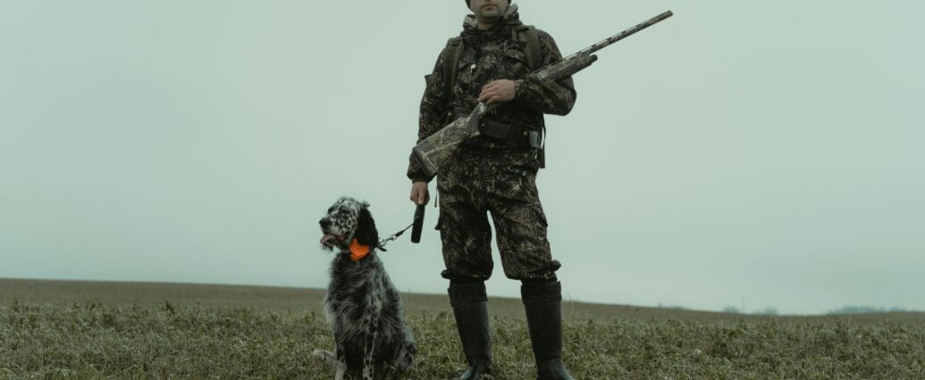 Man in camouflage with hunting dog and rifle in winter field.