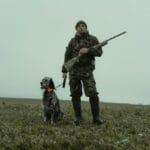 Man in camouflage with hunting dog and rifle in winter field.