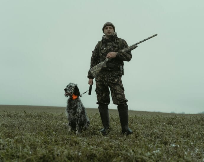 Man in camouflage with hunting dog and rifle in winter field.