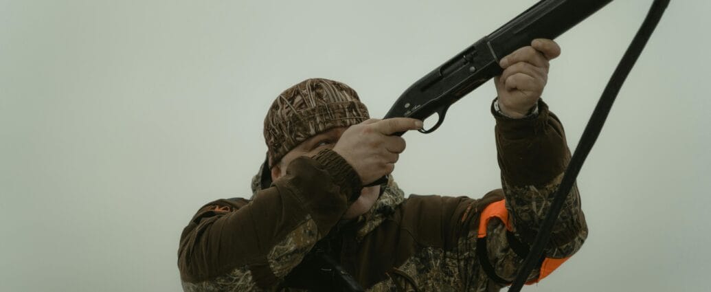 Hunter wearing camouflage gear aiming a shotgun in an open field setting, demonstrating outdoor hunting activity.