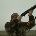 Hunter wearing camouflage gear aiming a shotgun in an open field setting, demonstrating outdoor hunting activity.