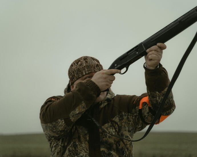 Hunter wearing camouflage gear aiming a shotgun in an open field setting, demonstrating outdoor hunting activity.