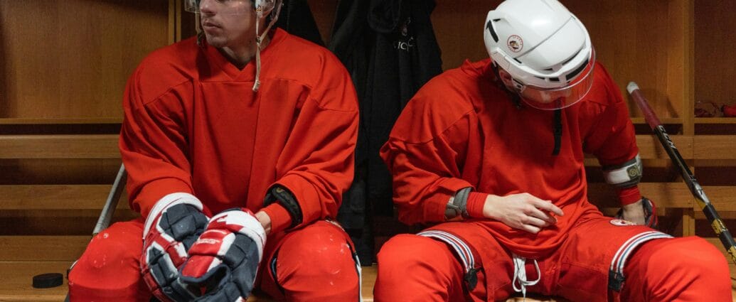 Two hockey players in red uniforms rest in the locker room.
