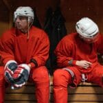 Two hockey players in red uniforms rest in the locker room.