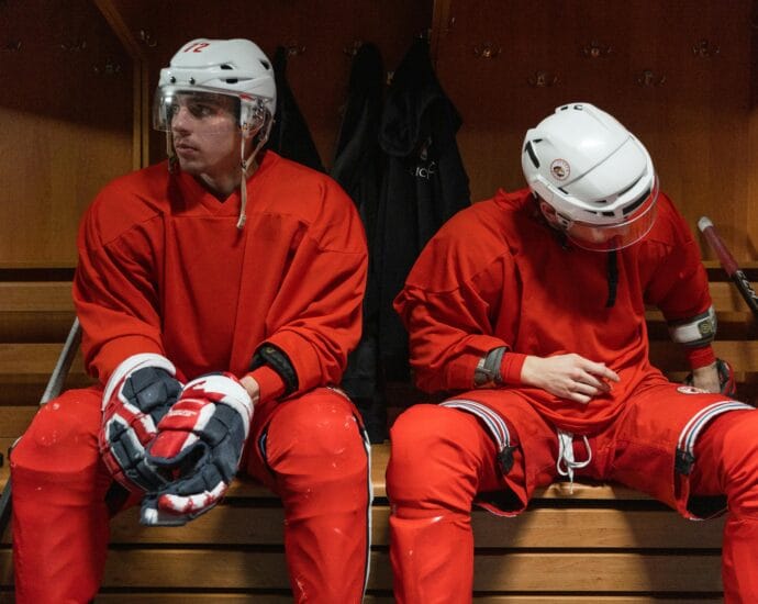 Two hockey players in red uniforms rest in the locker room.