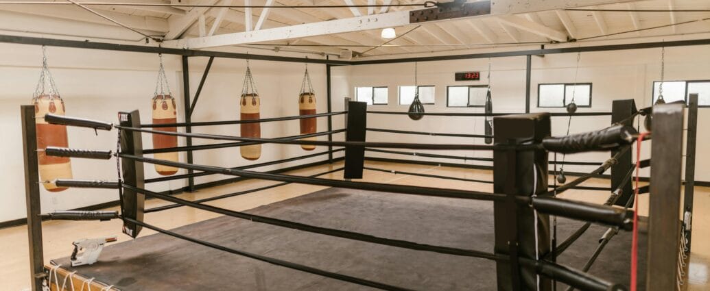 Interior view of a spacious boxing gym featuring a central ring and multiple heavy punching bags.