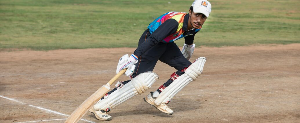 A cricket player in colorful attire bats during a daytime match on a sunny field.