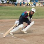 A cricket player in colorful attire bats during a daytime match on a sunny field.