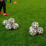 Pile of soccer balls on a green field with a person preparing for training session.