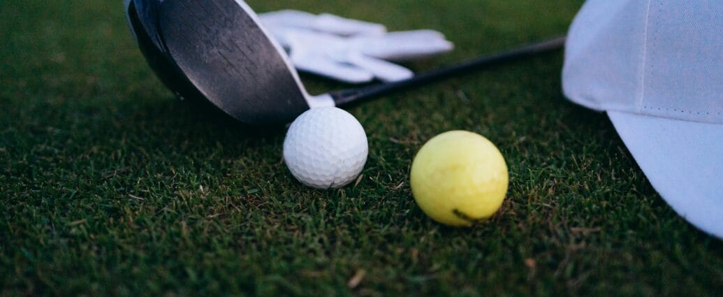 Close-up shot of a golf club, golf balls, and a cap on grass.