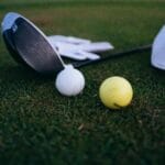 Close-up shot of a golf club, golf balls, and a cap on grass.